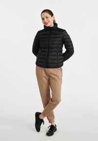 Czarna kurtka puchowa z wysokim kołnierzem i zapięciem na zamek, w połączeniu z luźnymi beżowymi spodniami i czarnymi sneakersami. Gładka tekstura, minimalistyczny design.