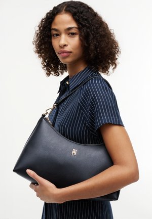 Jonge vrouw met krullend haar die een navyblauwe jurk met krijtstrepen draagt, en een zwarte leren handtas met gouden logo en schouderriem vasthoudt.