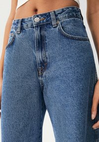 Jeans de denim azul de cintura alta con un acabado desgastado, con costuras naranjas, cinco bolsillos y un cierre de botón metálico en la cinturilla.