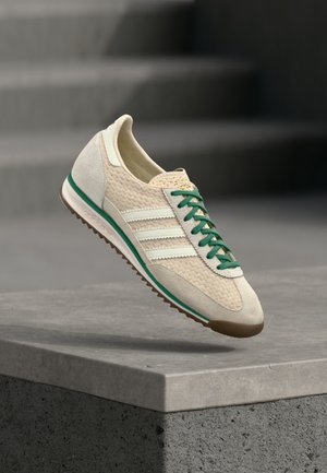 Beige sneaker med grønne snørebånd, hvide striber og brun sål, der svæver over en betonforkant med slørede grå trapper i baggrunden.