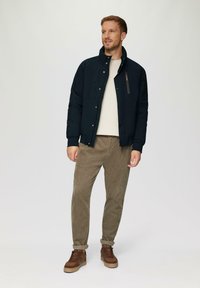 Marineblauwe pufferjack met een hoge kraag, drukknoopsluiting en zijzakken, gecombineerd met een lichtbeige sweater en een paarse corduroy broek.