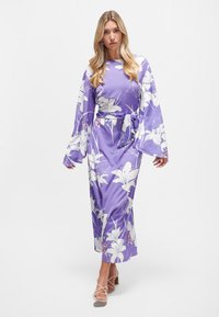 Langärmliges, lila Blumenkleid mit weißen Lilien und Akzenten. Aus glattem Stoff gefertigt, mit einem taillierten Gürtel und fließenden Ärmeln.