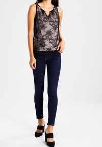 Top in pizzo nero con dettagli a occhiello sul collo, abbinato a jeans skinny blu scuro e scarpe con tacco a blocco nere.