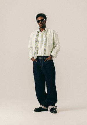 Homme portant des lunettes de soleil, chemise blanche ample à rayures avec cravate à motifs dénouée, jean large foncé retroussé aux chevilles et chaussures noires sur fond uni.