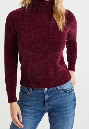Femme portant un pull ajusté à col roulé bordeaux à manches longues avec un jean bleu sur un fond uni.