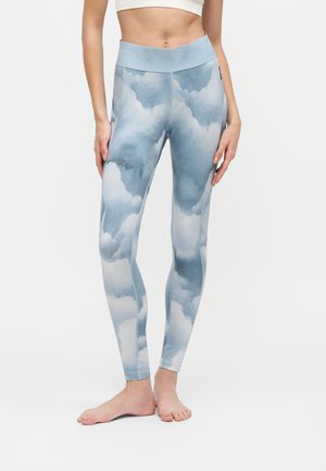 Femme portant un legging bleu taille haute avec un motif nuage, debout pieds nus sur un fond clair uni.
