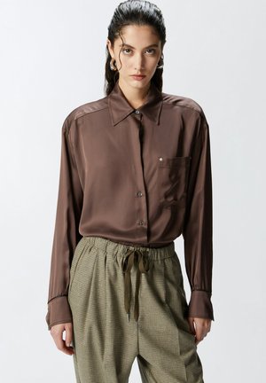 Camicia di raso marrone con colletto a punta, taschino frontale e chiusura con bottoni. Abbinata a pantaloni a quadri verdi con vita elastica.
