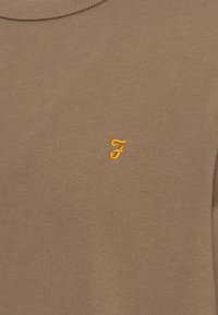 Tessuto a maglia marrone con piccola lettera "F" ricamata in corsivo giallo, centrata vicino alla parte alta.