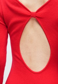 Czerwony dziergany top z długimi rękawami, ozdobą w postaci kokardki z tyłu oraz dużym okrągłym wycięciem poniżej linii szyi.