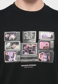 Sort bomulds t-shirt med et grafisk motiv af flere vintage fjernsyn, der viser forskellige billeder, med teksten "WEEKEND OFFENDER" nedenfor.