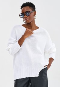 Biały, oversize'owy sweter dziewiarski z ribbowym wykończeniem i szerokimi rękawami, zestawiony z dużymi okularami w kształcie żółwia i kolczykami w kształcie kół.