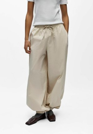 Personne portant un pantalon large beige à taille coulissante avec ourlets élastiques, un haut blanc à manches courtes, des chaussettes grises et des chaussures marron foncé à bouts carrés.