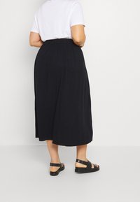 Persona che indossa una maglietta bianca a maniche corte infilata in una gonna midi nera a vita alta e sandali neri con cinturini, in piedi contro uno sfondo semplice.