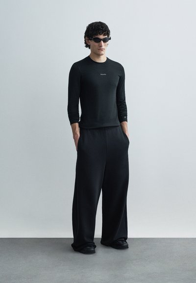 Black long-sleeve shirt with logo, loose black trousers, and black sunglasses. Smooth texture and relaxed fit. Worn by a model in a minimal setting.