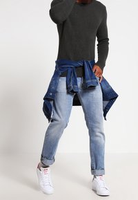 Grau strukturiertes Strickpullover, blaue Jeans mit hochgerollten Säumen und weiße Sneakers. Blaue Jeansjacke um die Taille gebunden.