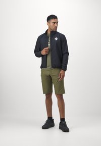 Svart lett jakke med glidelås, elastiske mansjetter og logo; grønn t-skjorte med geometrisk mønster; olivengrønne shorts; svarte sko.