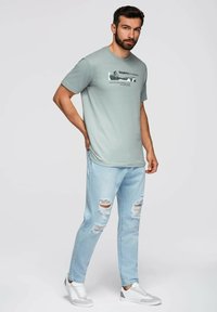 Camiseta de algodón verde claro con mangas cortas que presenta un gráfico impreso en negro. Combinada con unos vaqueros desgastados de color azul claro y zapatillas blancas.