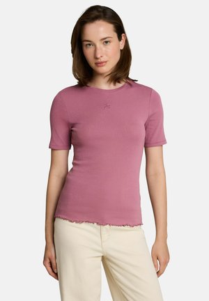 Jonge vrouw met een aansluitend stofroze kort mouwen topje met gegolfde zoom en lichte beige broek, staand tegen een effen achtergrond.