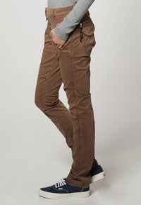 Person wearing brown slim-fit pants with button fly, side zipper pocket, and navy blue sneakers, hand in pocket, standing on gray background.