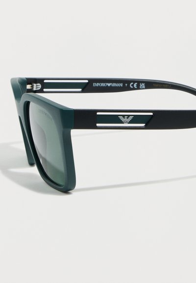 Μαύρα και σκουροπράσινα τετράγωνα γυαλιά ηλίου με χοντρά πλαίσια και λογότυπο Emporio Armani στο μπράτσο του βραχίονα, πάνω σε λευκό φόντο.