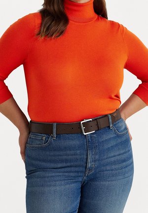 Kvinde iført en lys orange rullekravesweater stoppet ned i blå jeans med et brunt bælte, hænderne hvilende på hofterne.