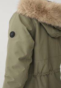 Parka verde oliva con un borde de capucha de pelaje sintético, cintura elástica y un parche redondo negro con el logo en la manga izquierda. Textura de tejido suave.