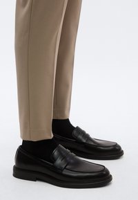 Μαύρα δερμάτινα μοκασίνια με οριζόντιες ραφές, συνδυασμένα με μπεζ καλοραμμένο παντελόνι και μαύρες κάλτσες, σε ουδέτερο φόντο.