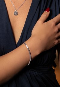 Bracelet jonc de couleur argentée avec une finition lisse et polie, porté sur un bras bronzé, sur un fond de tissu bleu foncé.