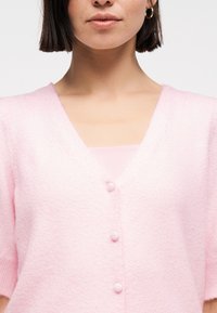 Γυναίκα με κοντά σκουρόχρωμα μαλλιά, που φοράει χρυσούς κρίκους σκουλαρίκια και ροζ ανοιχτόχρωμο αφράτο ζακετάκι με τρία κουμπιά καλυμμένα από ύφασμα.