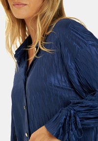 Blusa texturizada azul marino con cierre de botones, cuello y detalles de mangas fruncidas. La tela tiene un brillo sutil y un diseño plisado.