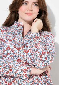 Camisa floral con un cuello alto, que presenta patrones rojos y azules sobre un fondo blanco. Tejido suave con detalles de botones en los puños.