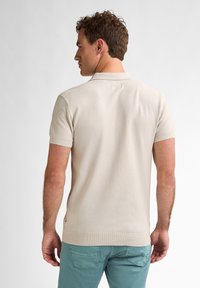 Beige polo-shirt med korte ærmer, struktureret mønster, ribbet krave og manchetter, let figursyet design, båret med teal-farvede bukser.