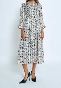 Robe longue à motifs avec col cranté, boutonnage avant et manches cloches. Couleurs incluant noir, blanc et pêche. Associée à des talons noirs.