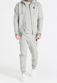 Felpa grigia con cerniera e coulisse, abbinata a pantaloni cargo in felpa coordinati, dotati di tasche e dettagli logo sottili. Sneakers bianche completano il look.