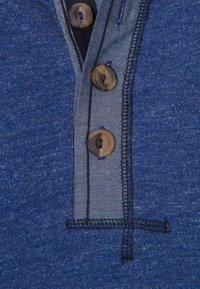 Polo blu con tessuto testurizzato, tre bottoni in materiale marrone chiaro e colletto a contrasto in blu navy. Dettagli di cucitura visibili.