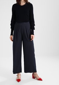 Black sweater with layered ruffle sleeves, paired with high-waisted navy wide-leg trousers and beige heels with red pointed toes.