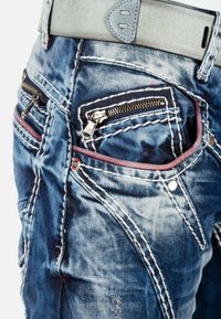 Jeans in denim con colore blu sbiadito, dotati di tasche con doppia cerniera, cuciture rosse a contrasto e una cintura in pelle grigia con finitura liscia.