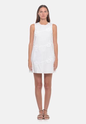 Donna che indossa un miniabito bianco senza maniche con ricamo floreale e sandali con cinturini e grandi perle, in piedi su uno sfondo bianco.