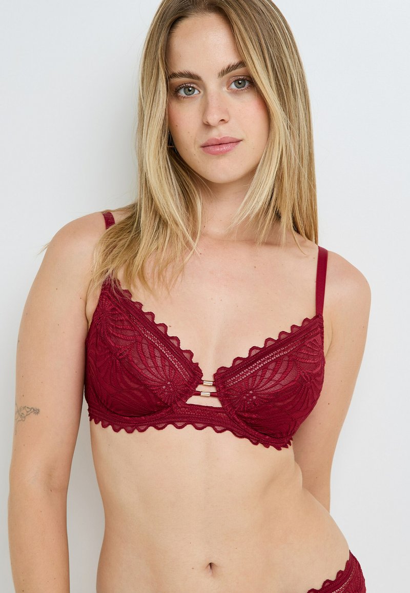 Reggiseno in pizzo bordeaux con bordi ondulati, scollatura a V e decorazioni dorate. Presenta un delicato motivo floreale. Spalline regolabili.