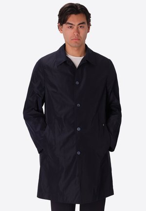 Uomo con cappotto nero abbottonato e mani nelle tasche, in piedi davanti a uno sfondo chiaro e uniforme.