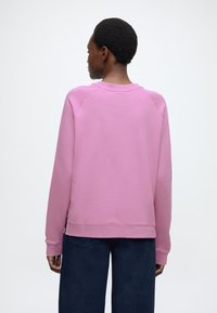 Rozā džemperis ar apaļu kakla izgriezumu, ribota apdare piedurknēm un apakšmalai. Raksturots ar brīvu piegriezumu un gludu tekstūru. Valkāts kopā ar tumši ziliem platajiem džinsiem.