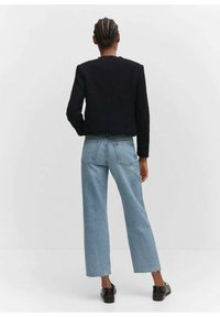 Giacca nera texturizzata abbinata a jeans a gamba larga blu chiaro, dotati di tasche posteriori. Il modello si trova su uno sfondo neutro, con la schiena rivolta verso la fotocamera.