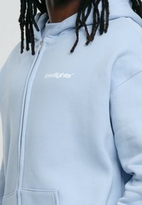 Sweat à capuche bleu clair zippé en tissu doux, avec une poche kangourou et un petit logo blanc sur la poitrine. Capuche avec cordon de serrage.