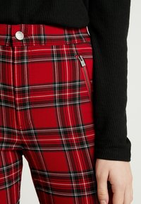 Pantaloni a quadri rossi con linee nere e bianche, dotati di una zip laterale, abbinati a un top nero a maniche lunghe con texture.