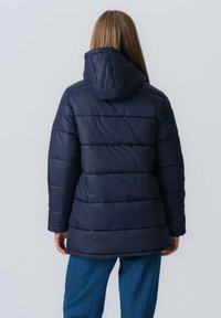 Marineblå pufferjakke med hætte, der har et quiltet design og lange ærmer. Materialet synes at være syntetisk og en smule skinnende.