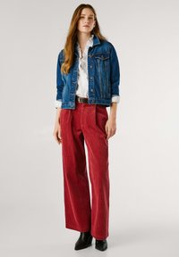 Giacca di jeans, camicia bianca, pantaloni larghi in corduroy rosso, stivaletti neri alla caviglia. Presenta cuciture leggere, dettagli delle tasche e tessuto testurizzato.