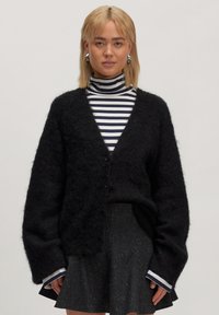 Schwarze, flauschige Strickjacke mit tiefem V-Ausschnitt, ausgestattet mit zwei Knöpfen, getragen über einem schwarz-weißen gestreiften Rollkragenpullover und einem glitzernden Rock.