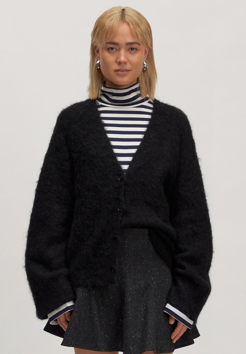 Schwarze, flauschige Strickjacke mit tiefem V-Ausschnitt, ausgestattet mit zwei Knöpfen, getragen über einem schwarz-weißen gestreiften Rollkragenpullover und einem glitzernden Rock.