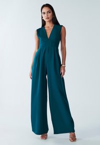 Türkisfarbener Overall mit tiefem V-Ausschnitt, eng anliegendem Oberteil und weitem Bein. Glatter Stoff mit minimalen Nähten und einem taillierten Detail.