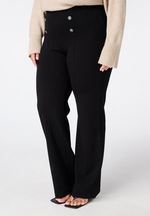 Vrouw draagt zwarte hoge taille broek met zilveren knopen en beige lange mouwen trui, staat met voeten in zwarte open-toe hakken.